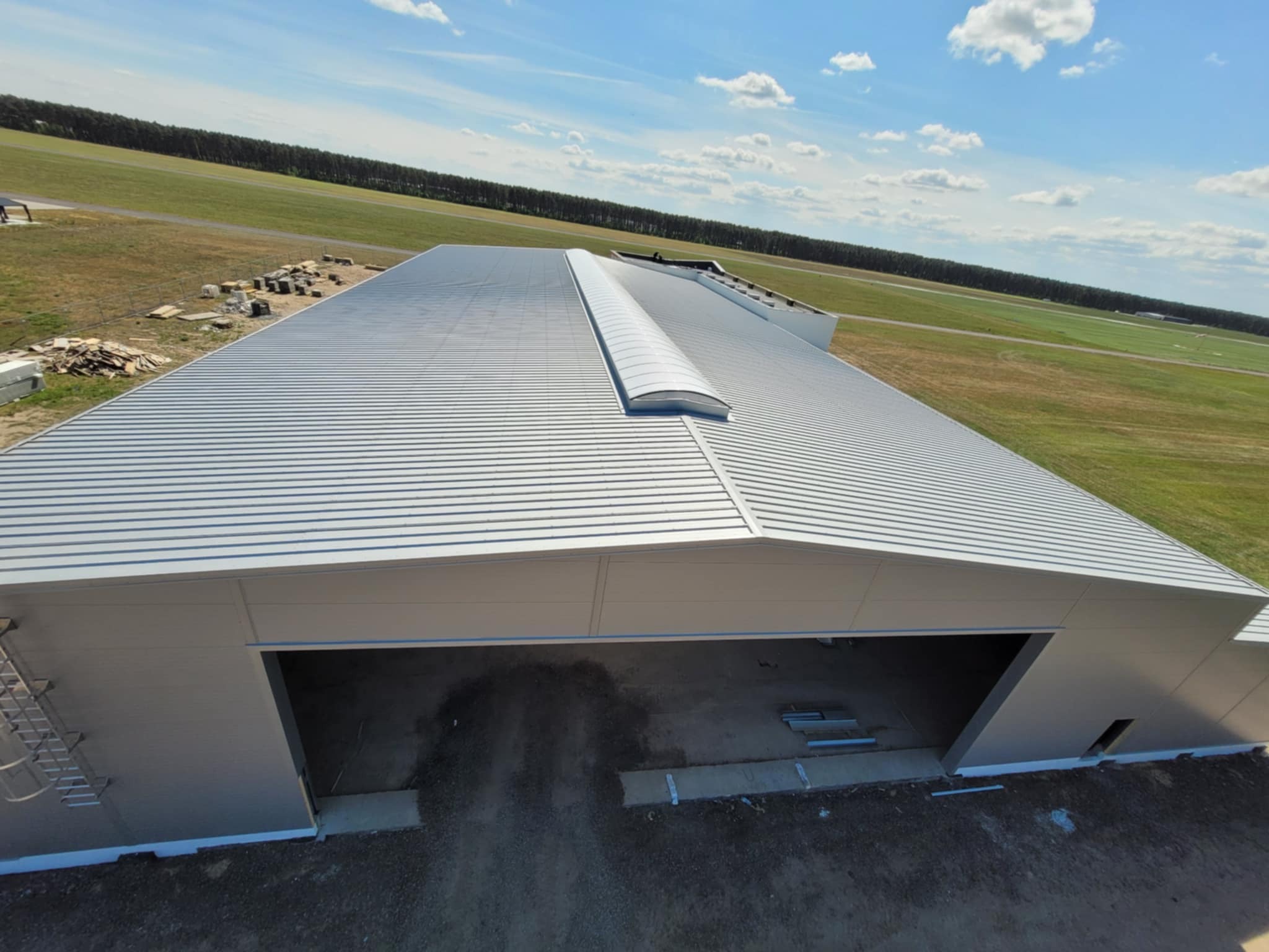 Zrealizowana hala stalowa z dachem z płyt warstwowych, widok z góry, błękitne niebo w tle. Widoczny fragment lotniska.