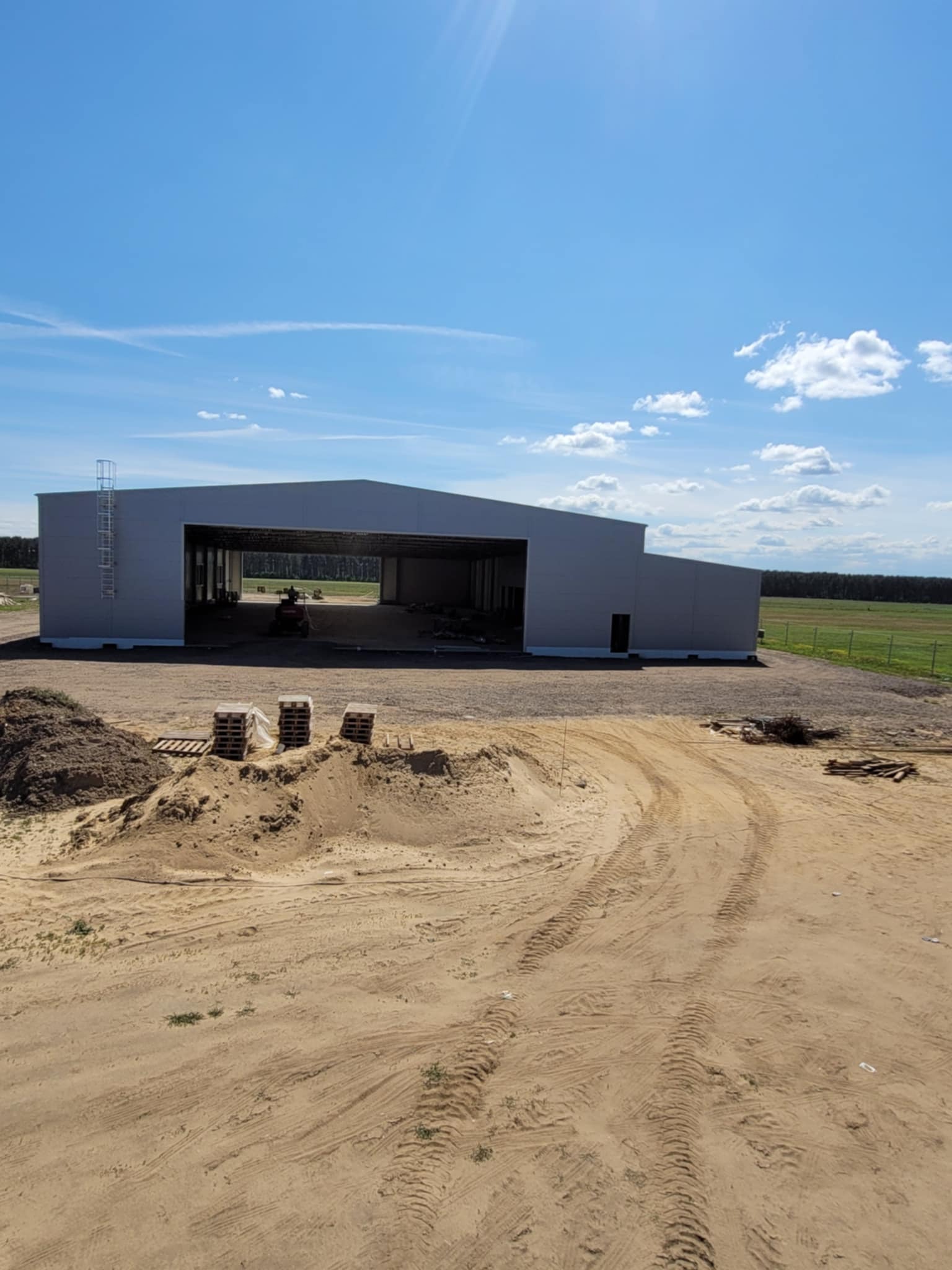 Konstrukcja hali stalowej w trakcie budowy, widok z zewnątrz na tle błękitnego nieba. Na pierwszym planie piasek ze śladami opon i palety.
