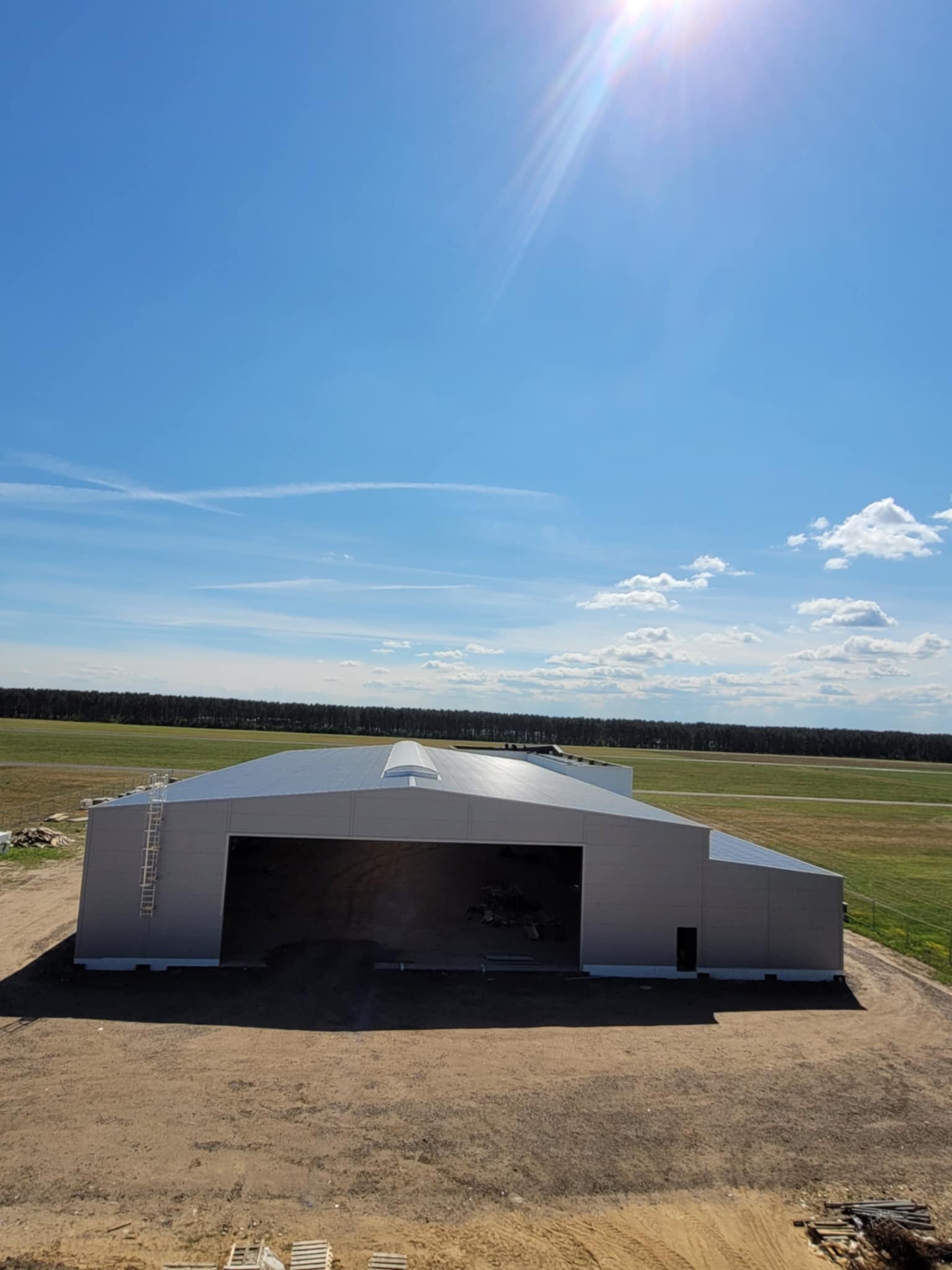Gotowa hala magazynowa z szarej płyty warstwowej na tle zielonych łąk i błękitnego nieba z chmurami. Widoczna drabina na bocznej ścianie i świetlik dachowy.