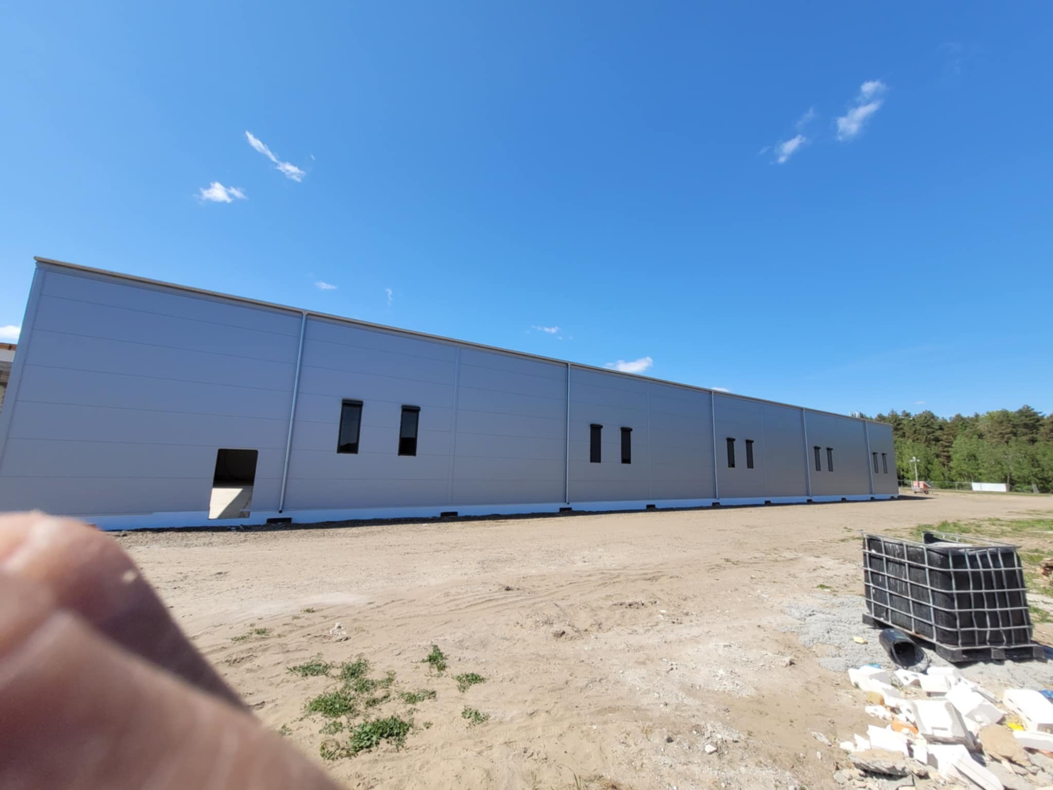 Szara hala z płyty warstwowej w trakcie budowy, widoczne okna i otwarty wjazd. Na pierwszym planie gruz i paletopojemnik. Błękitne niebo w tle.