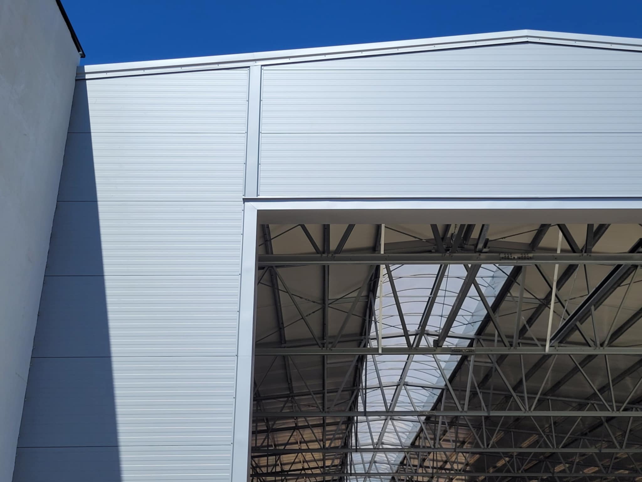 Fragment hali z płyt warstwowych, widok z zewnątrz na konstrukcję dachu przez otwór. Jasnoszare panele ścienne i stalowa konstrukcja pod błękitnym niebem.