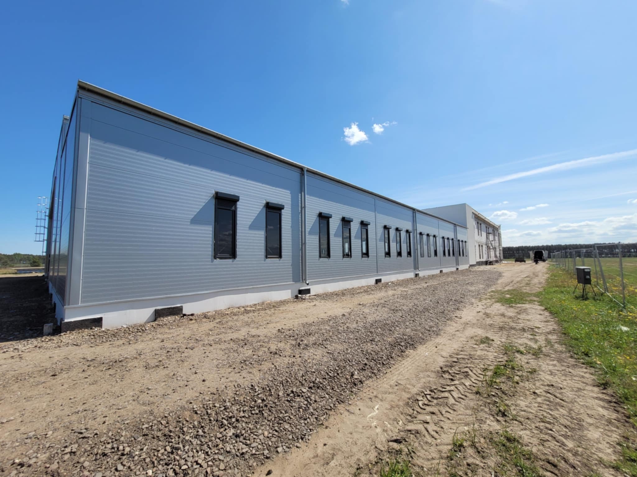 Nowoczesna hala z szarej płyty warstwowej, czarne okna, jasne niebo, teren w trakcie wyrównywania, widok z boku, w tle zieleń.