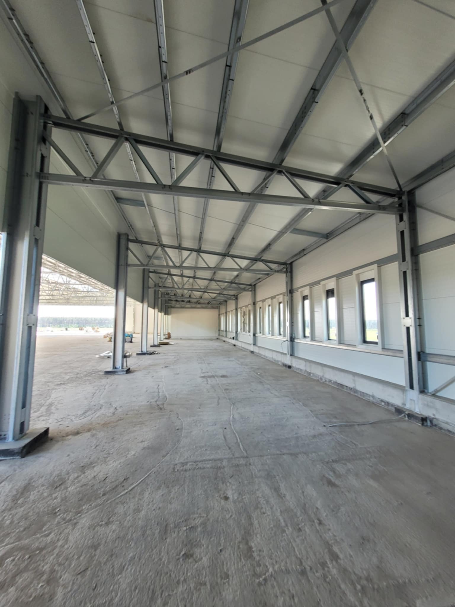 Wnętrze hali w trakcie budowy: stalowa konstrukcja, płyty warstwowe na dachu, rząd okien wzdłuż ściany. Widoczny surowy beton na podłodze i elementy konstrukcji na zewnątrz.