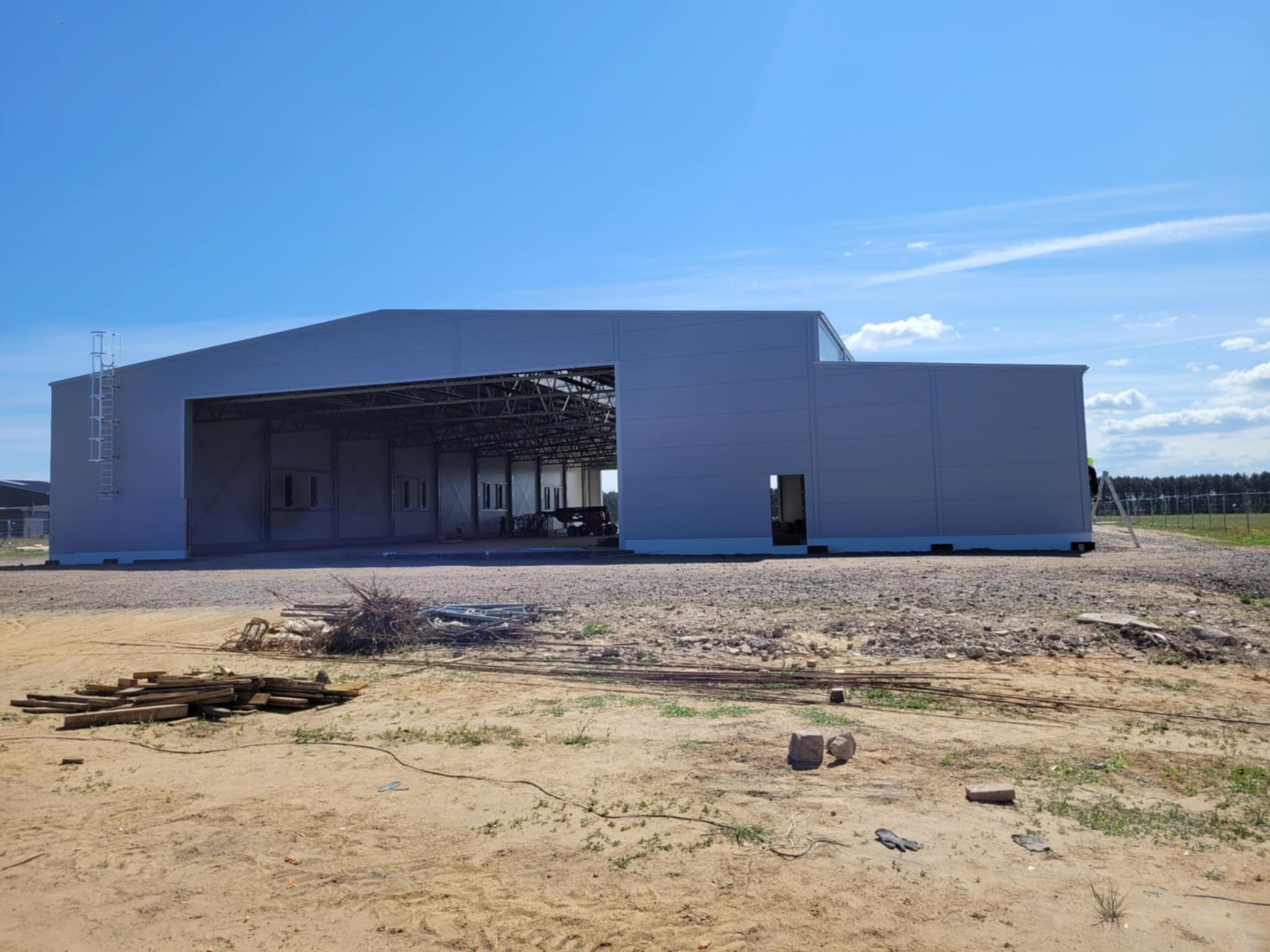Budowa hali z płyt warstwowych w Wierzchlas. Widok na gotową konstrukcję z szarej płyty, błękitne niebo w tle. Na pierwszym planie gruz i materiały budowlane.