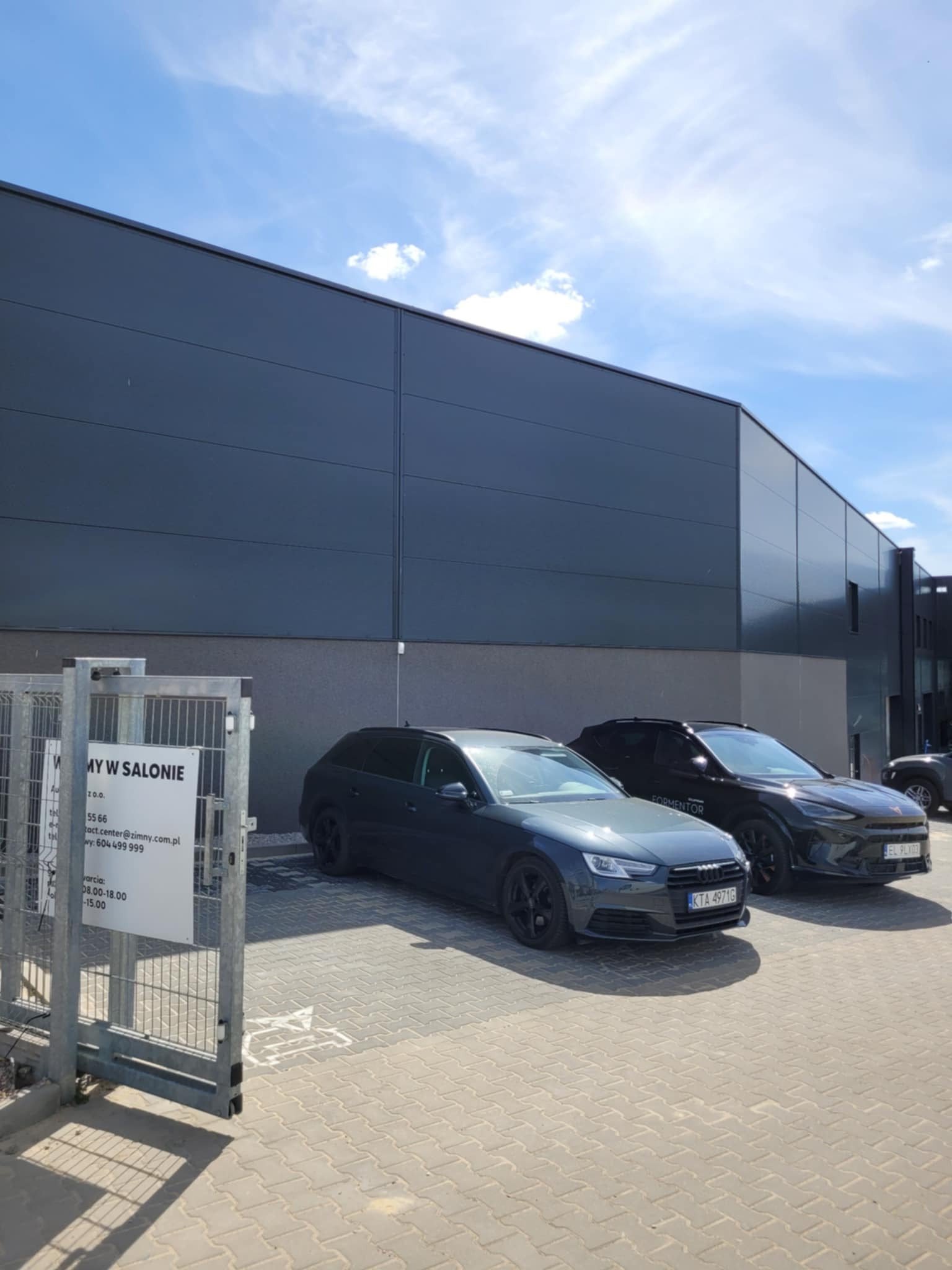 Nowoczesna hala z szarych płyt warstwowych, na tle błękitnego nieba. Przed budynkiem zaparkowane dwa samochody osobowe, w tym Audi A4 Avant. Widoczna brama wjazdowa.