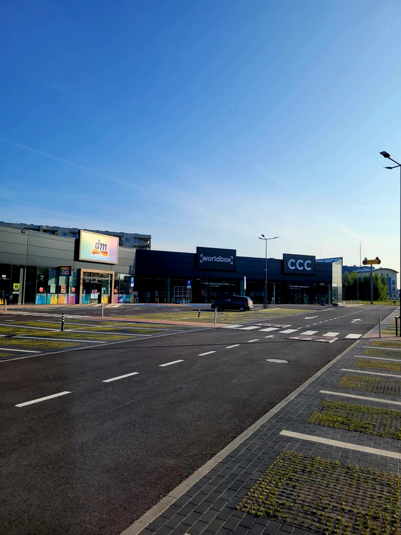 Nowoczesny kompleks handlowy z parkingiem i widocznymi szyldami sklepów DM, Worldbox i CCC w słoneczny dzień. Minimalistyczna architektura i czysta przestrzeń.
