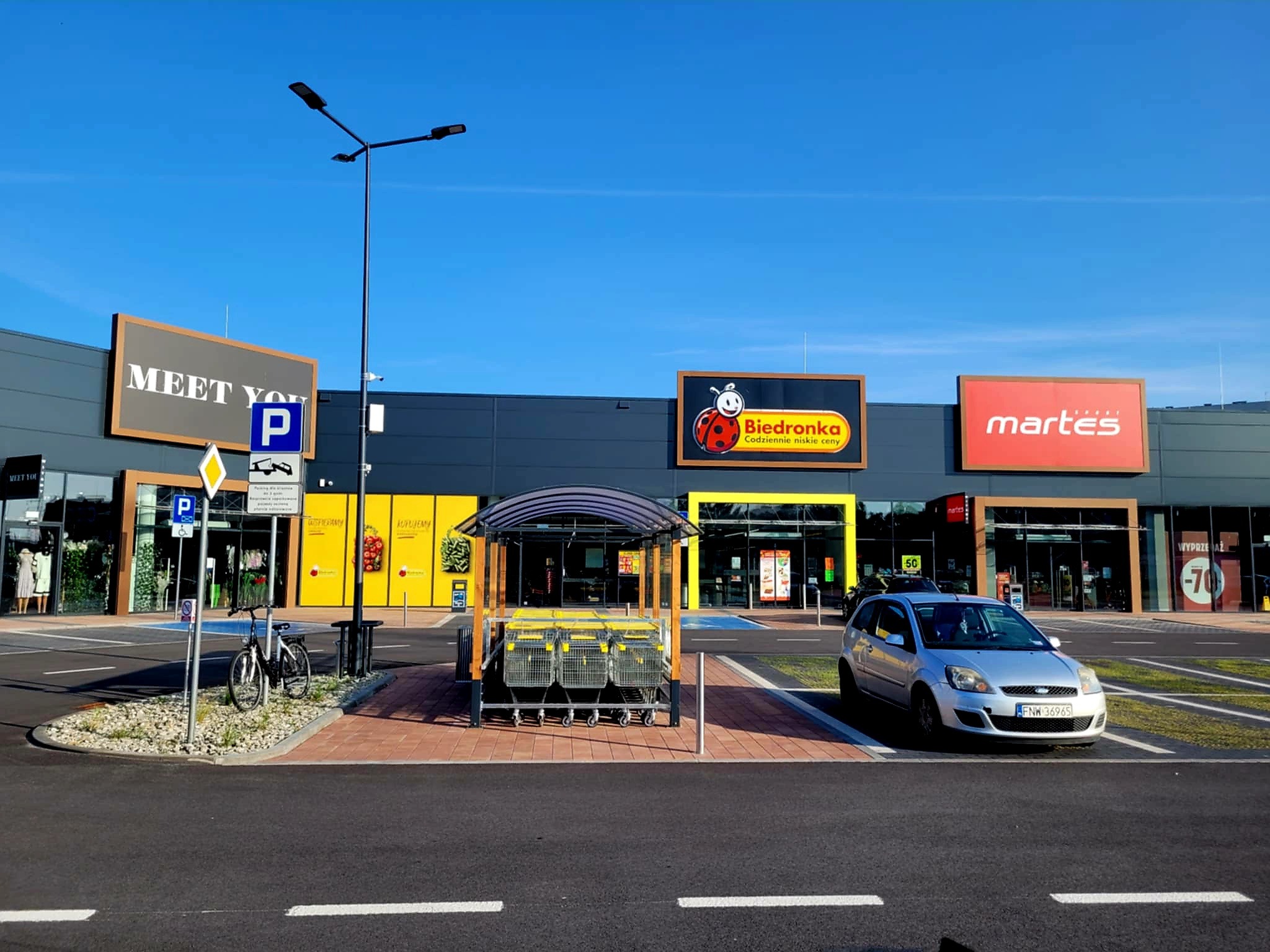 Hala handlowa z widocznymi szyldami sklepów: Biedronka, Martes i Meet You. Na pierwszym planie wózki sklepowe i zaparkowany samochód osobowy.
