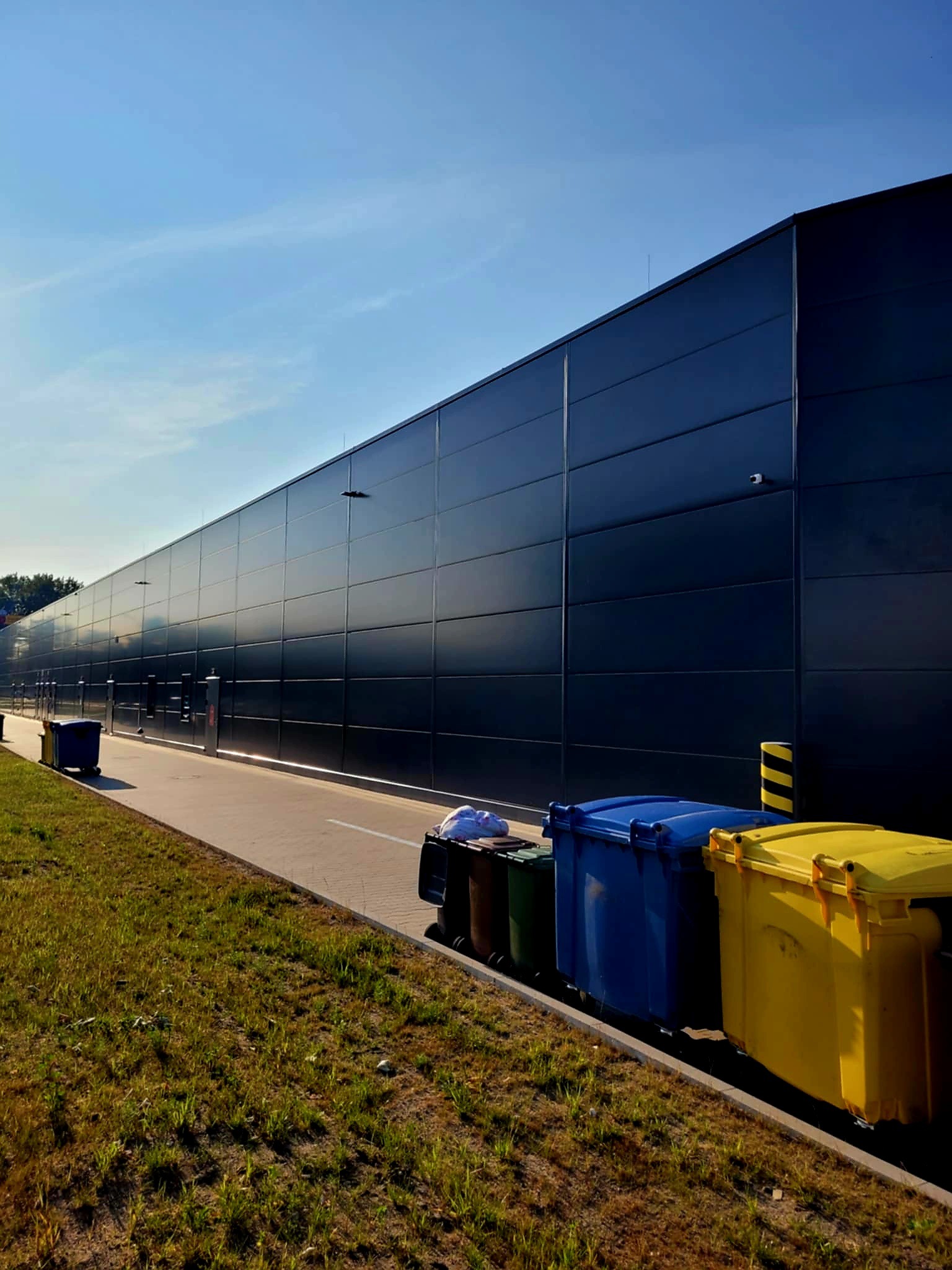 Nowoczesna hala z płyt warstwowych w odcieniach granatu, z widocznymi elementami zabezpieczeń i otoczeniem zieleni. Słoneczny dzień.