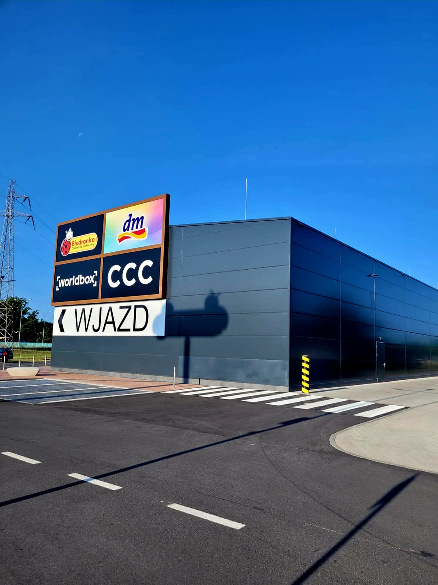 Nowoczesna hala z płyt warstwowych w odcieniach szarości, z dużym szyldem reklamowym Biedronki, DM, Worldbox i CCC nad wejściem. Widoczny wjazd na parking.