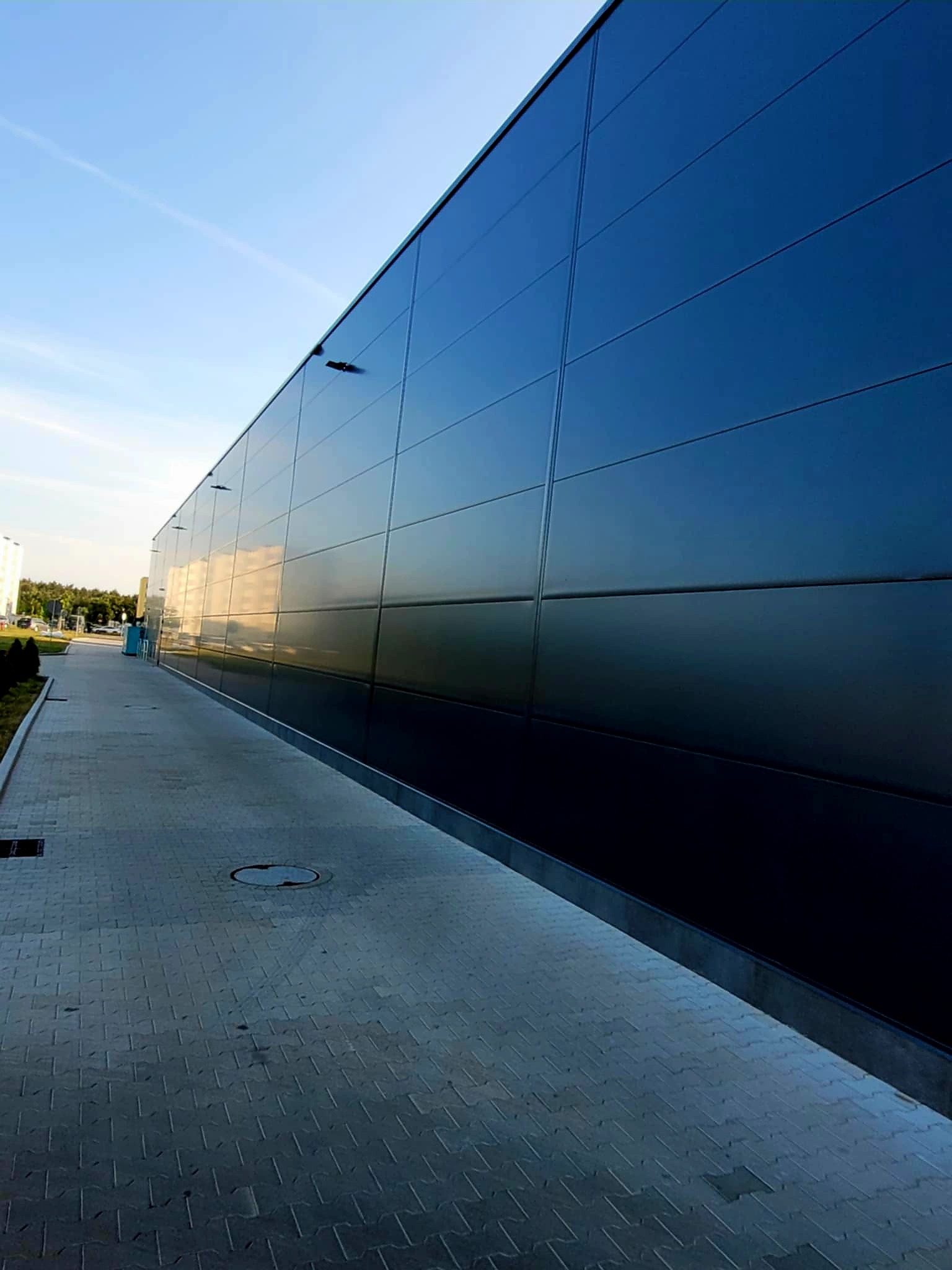 Ściana hali przemysłowej z paneli warstwowych, widok z dołu, z chodnikiem z kostki. Minimalistyczny design, odbicia nieba na panelach.