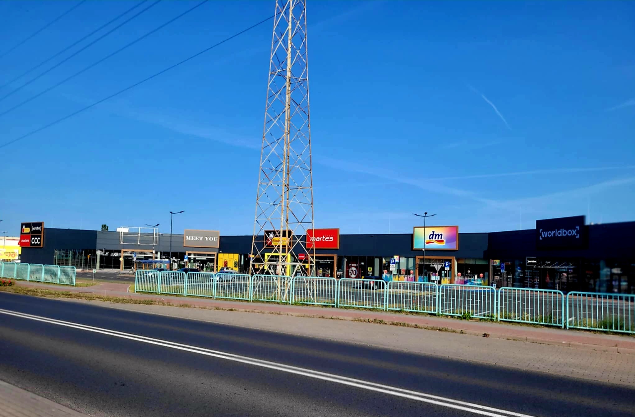 Nowoczesny kompleks handlowy z ciemną elewacją i widocznymi szyldami sklepów, ogrodzony metalowym płotem, z wysokim masztem energetycznym w tle.