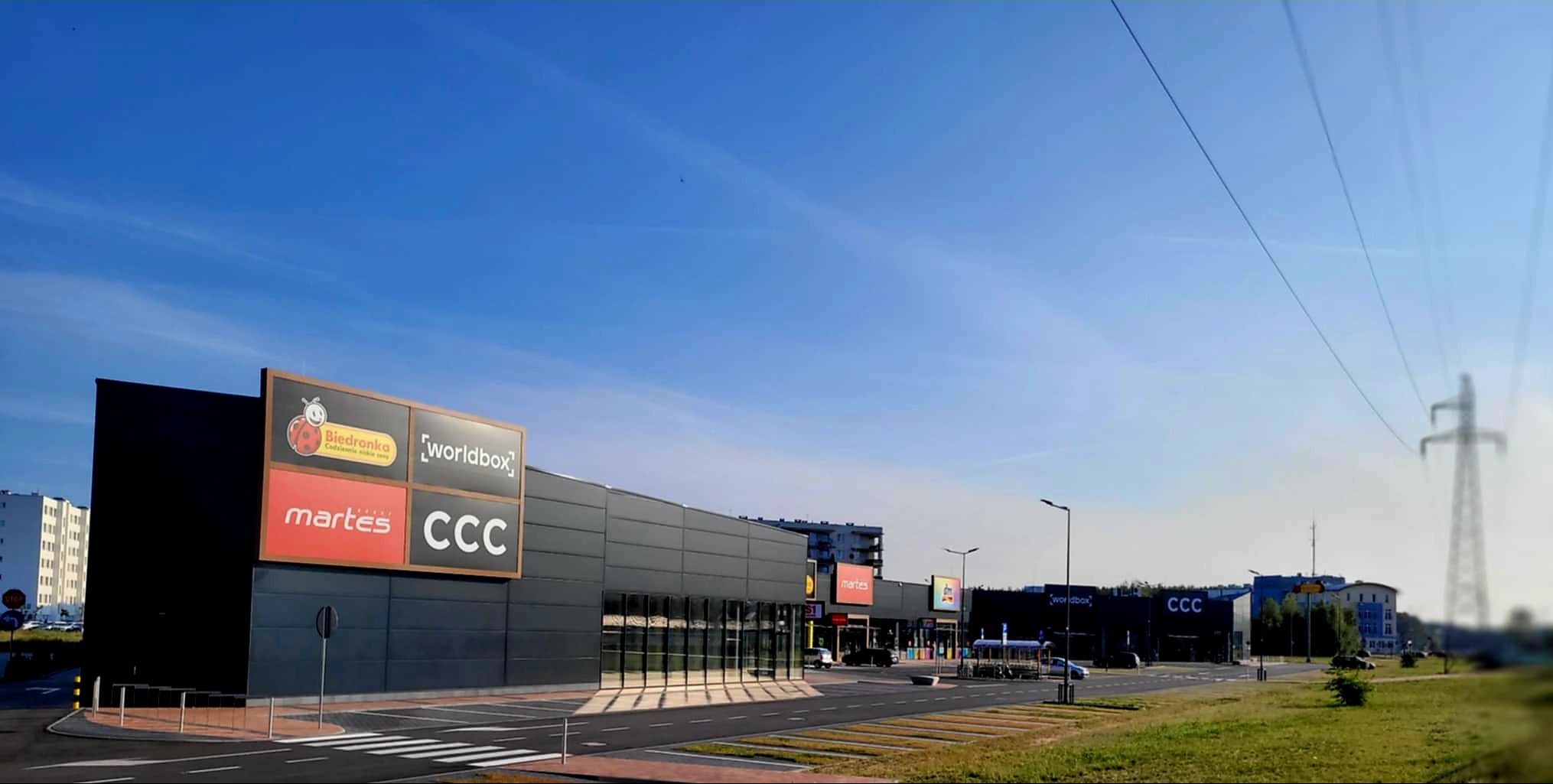 Nowoczesna hala handlowa z szarej płyty warstwowej, widok z zewnątrz w słoneczny dzień. Widoczne logo sklepów Biedronka, Martes, CCC, Worldbox.