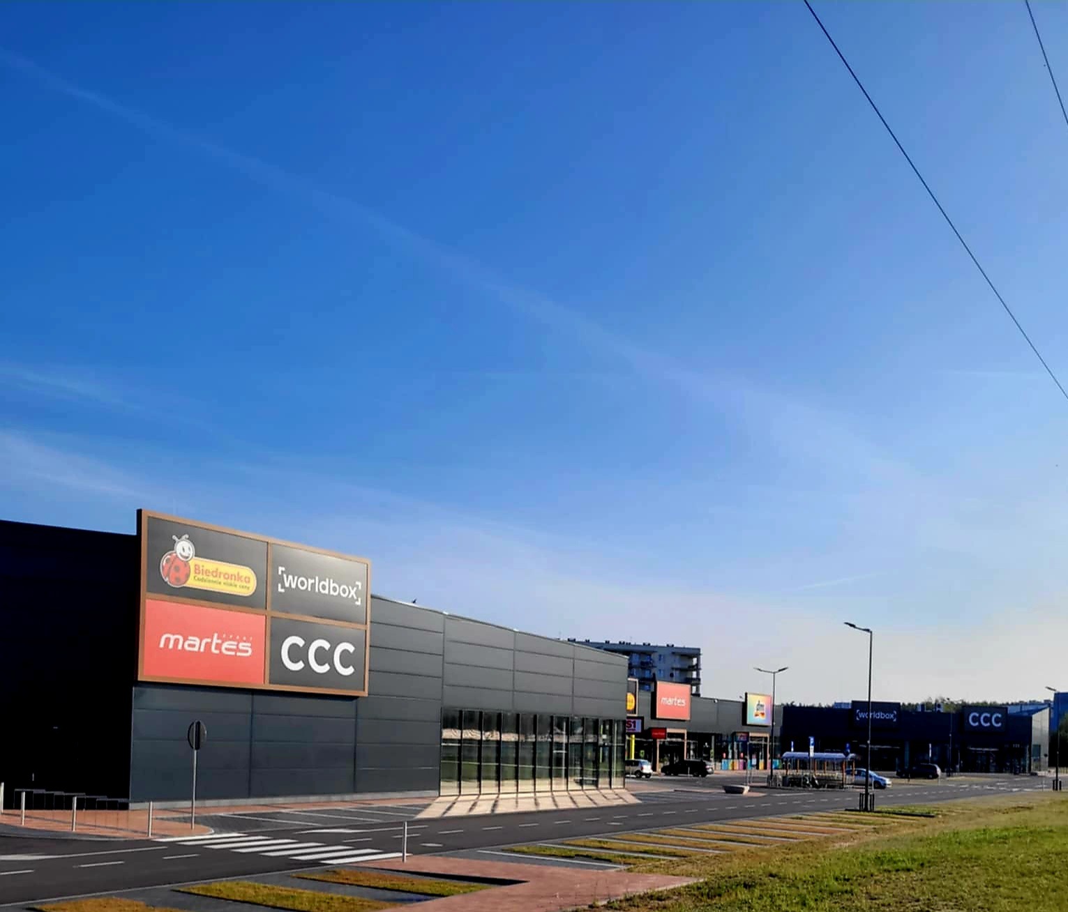 Nowoczesna hala handlowa z czarną elewacją z płyt warstwowych i logo Biedronka, Martes Sport i CCC, widziana z perspektywy ulicy w słoneczny dzień.