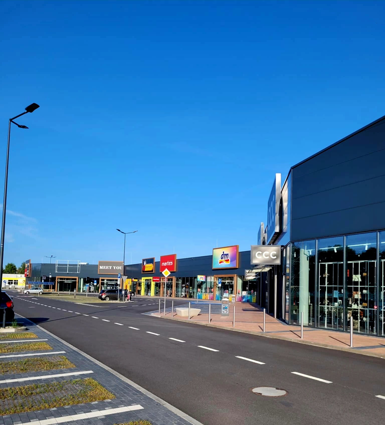 Nowoczesny budynek hali handlowej z płyt warstwowych, widok z zewnątrz, parkingu i drogi dojazdowej w słoneczny dzień. Widoczne logo sklepów.