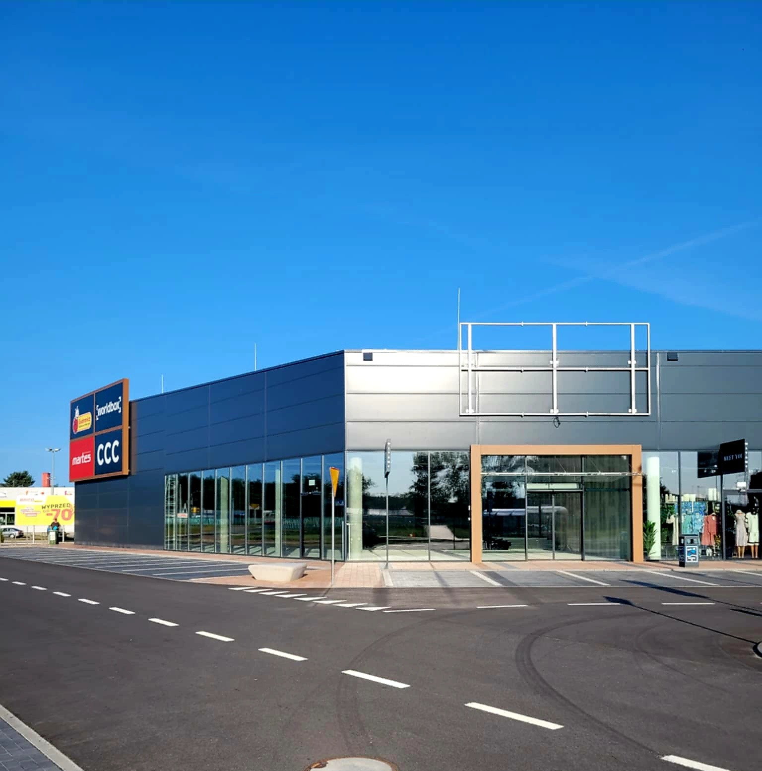 Nowoczesna hala handlowa z płyt warstwowych w kolorze szarym, z widocznymi witrynami sklepowymi i reklamami. Błękitne niebo w tle, asfaltowa droga na pierwszym planie.