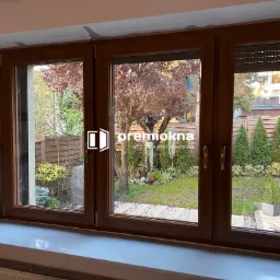 Trzyczęściowe okno z brązowymi ramami, widok na zadbany ogród z drzewami i ścieżką, logo firmy na szybie środkowego okna, biały parapet.