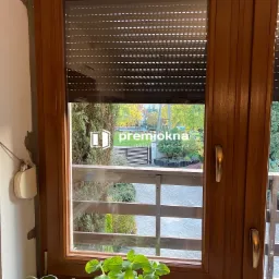 Brązowe okno z częściowo opuszczoną roletą zewnętrzną, widok na ogród zza szyby, doniczka z zieloną rośliną na parapecie wewnętrznym, logo firmy na szybie.