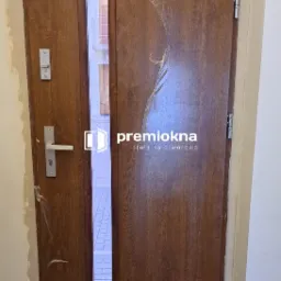 Brązowe drzwi wejściowe z małym oknem, częściowo zabezpieczone folią ochronną, widoczne ślady po montażu na ścianie, z logo firmy stolarskiej na środku.