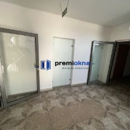 Wnętrze z dwoma otwartymi oknami aluminiowymi o szarych ramach, widoczne schody w tle jednego okna i ciemne pomieszczenie w drugim, na ścianie logo firmy stolarskiej, na podłodze brązowe płytki.