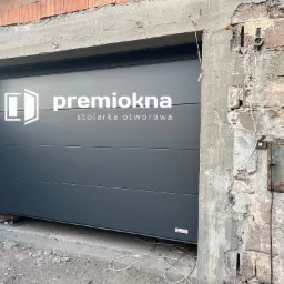 Szara segmentowa brama garażowa z logo 'premiokna stolarka otworowa' w surowym otoczeniu budowy, widoczne niedokończone mury i wystające przewody elektryczne.