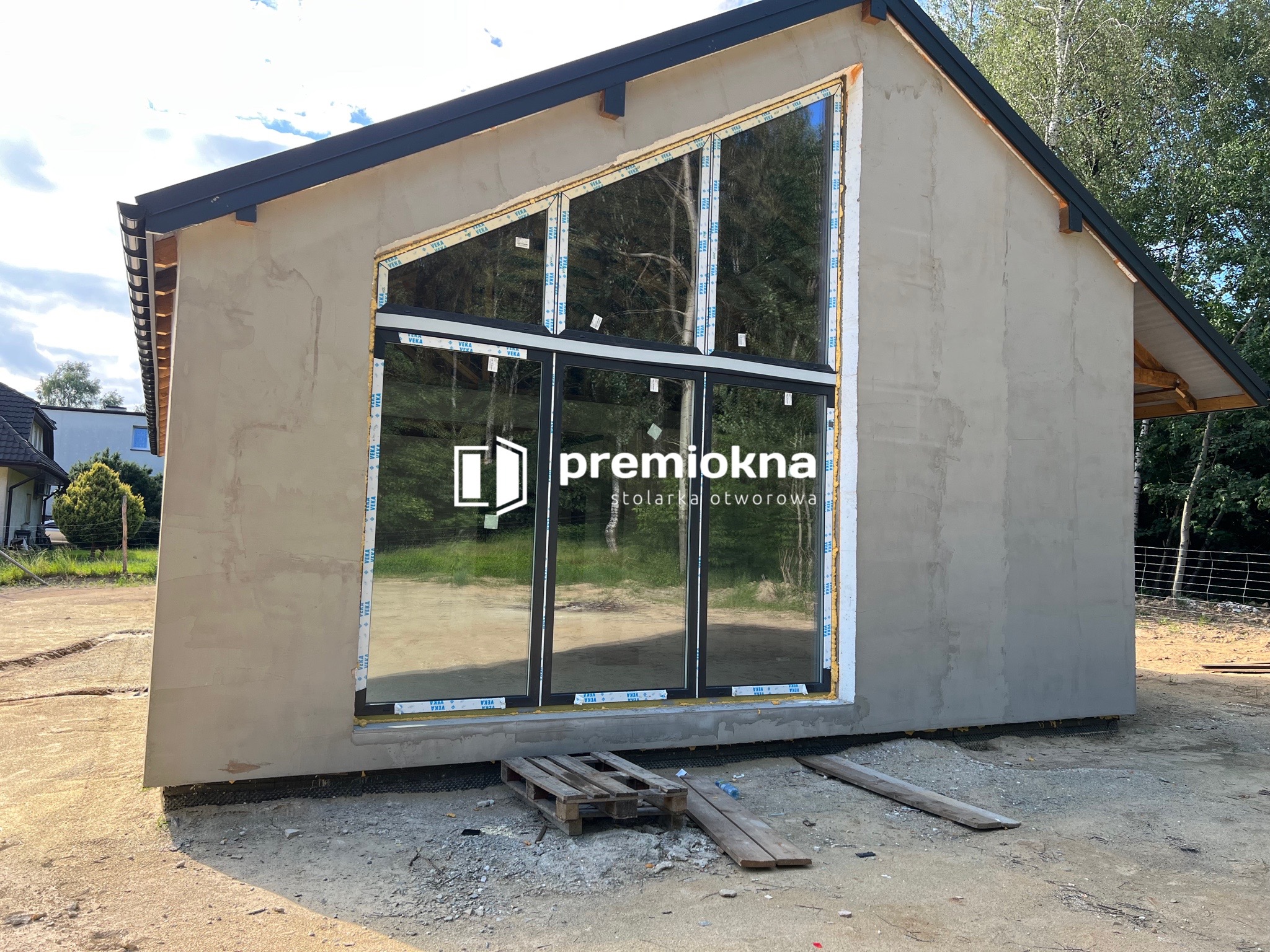 Nowo wstawione, duże okno o nietypowym, trójkątnym kształcie w surowym budynku, z widocznym logo producenta na szybie i drewnianą paletą pod oknem.