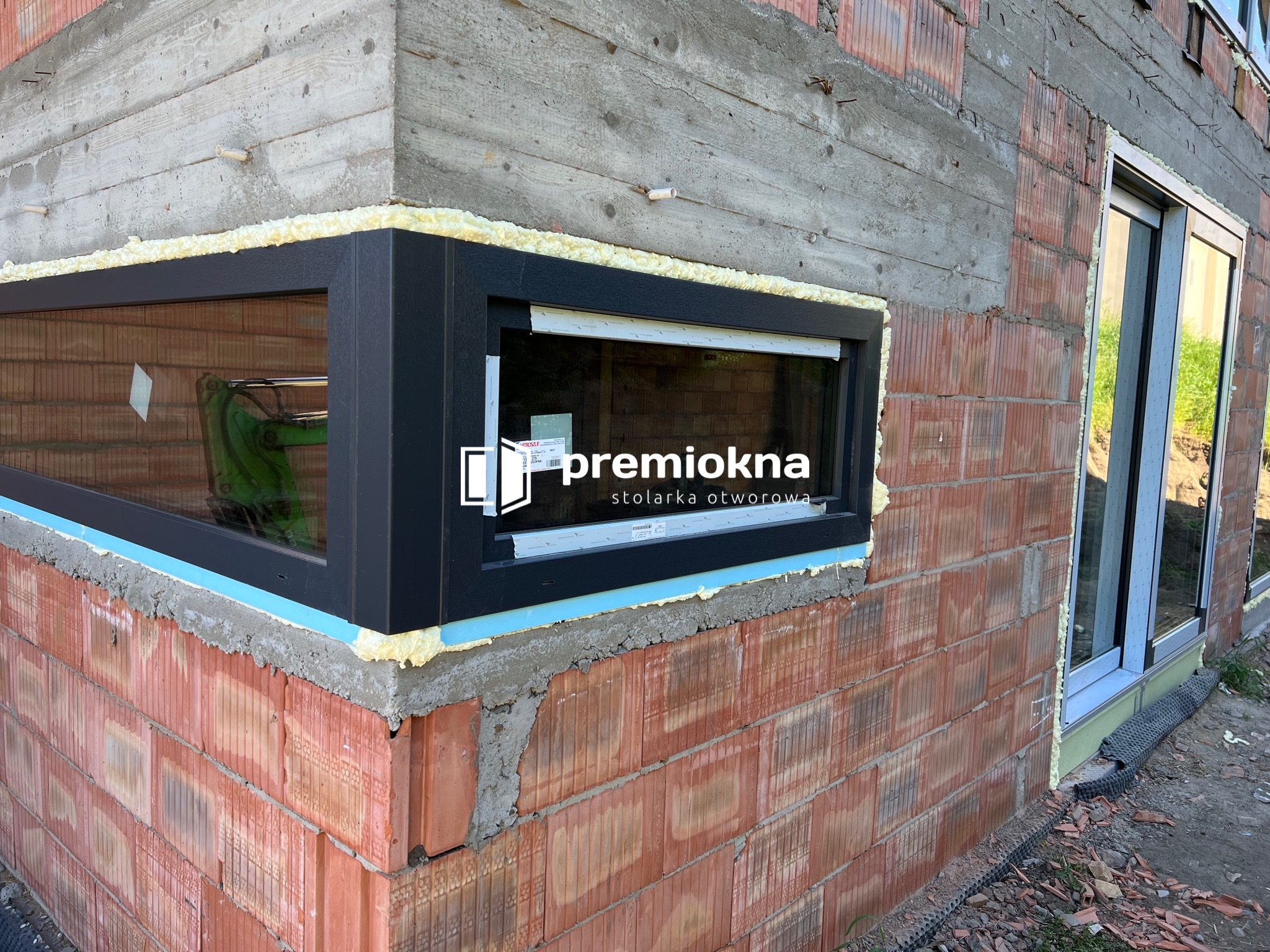 Montaż czarnego okna w surowym murze z cegły ceramicznej, pianka montażowa uszczelnia ramę, widoczny logotyp 'premiookna stolarka otworowa' na szybie.