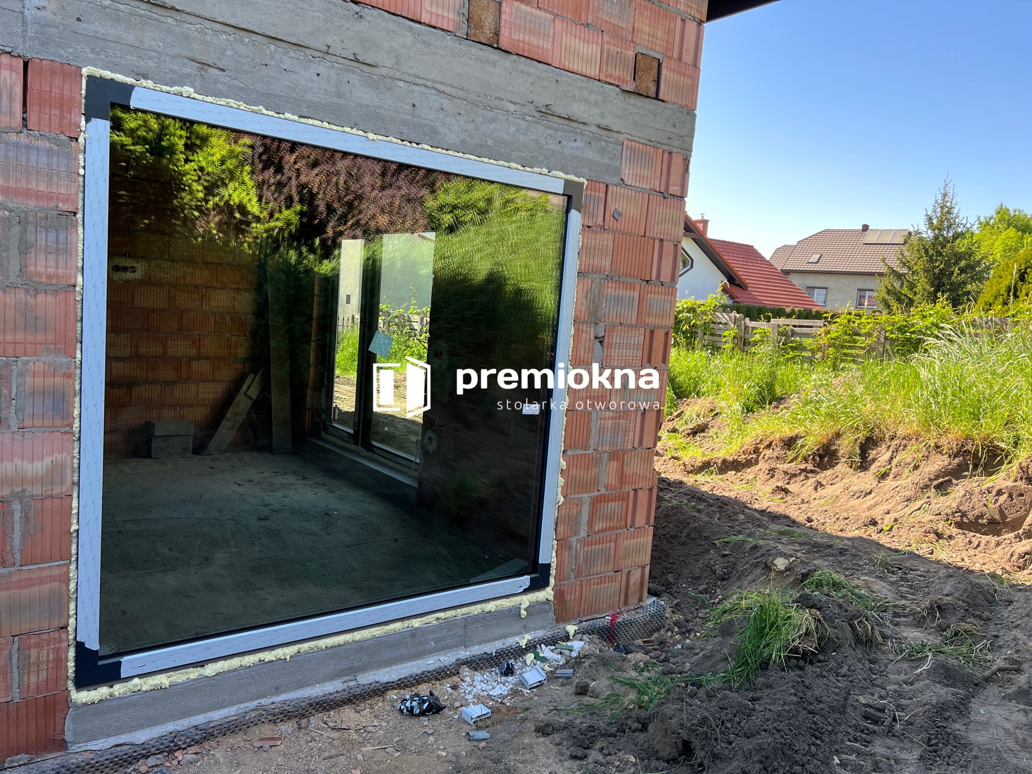 Montaż okna w surowym murze, widoczne wypełnienie pianką montażową, na szybie odbija się zieleń ogrodu, w prawym górnym rogu logo firmy.