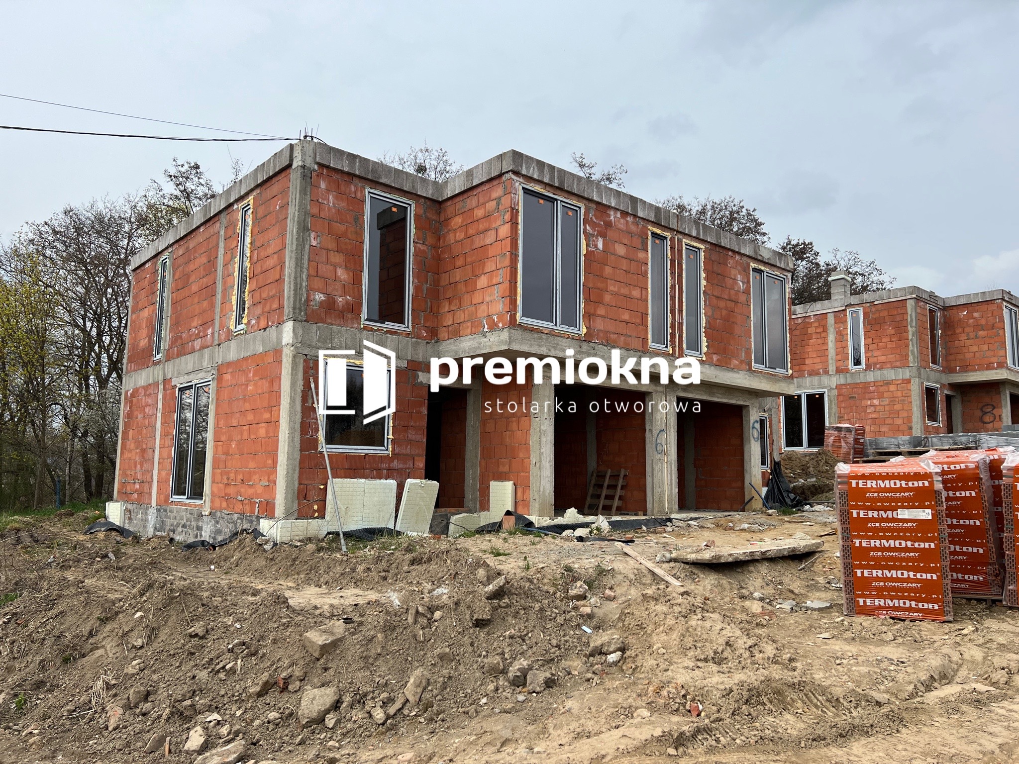 Surowy stan bliźniaka z częściowo zamontowanymi oknami z czarnymi ramami, widoczne palety z cegłami Termoton na placu budowy, logo firmy Premiokna na pierwszym planie.