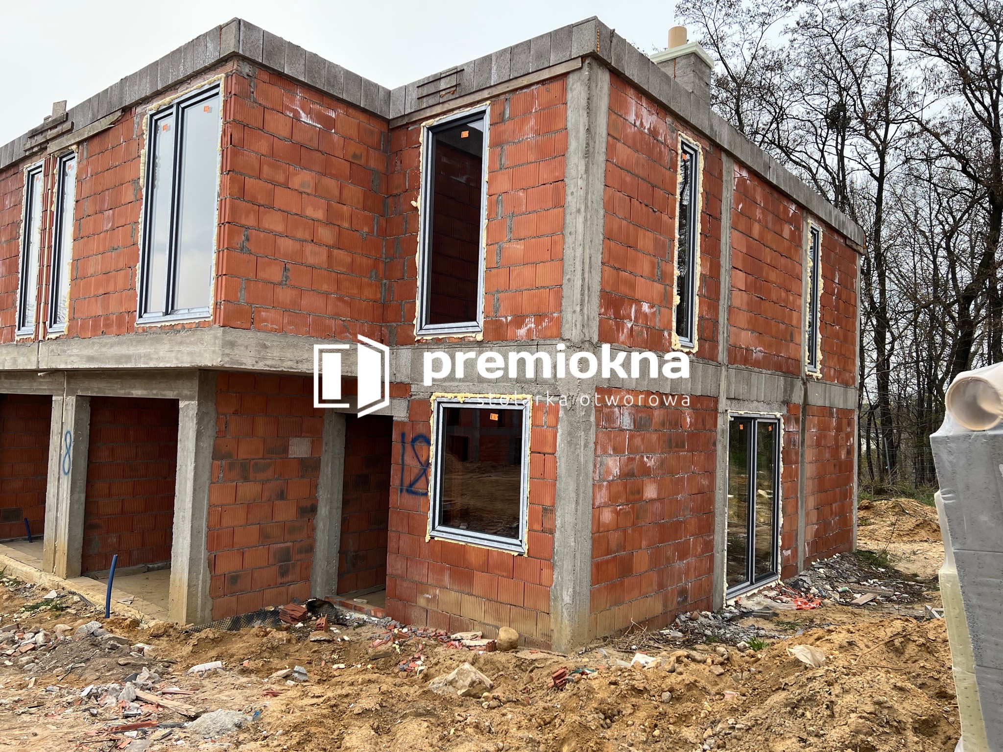 Surowy stan budynku w trakcie montażu okien z czarnymi ramami, widoczna pianka montażowa wokół okien, czerwona cegła i betonowe nadproża, teren budowy z ziemią i resztkami materiałów.