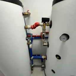 Dwa białe zbiorniki z widoczną instalacją hydrauliczną: rury miedziane, zawory i manometr. Instalacja w kolorach czerwonym i niebieskim, z detalami z mosiądzu.