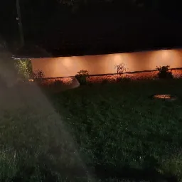 Nocne podlewanie trawnika zraszaczy w ogrodzie, oświetlony budynek w tle.