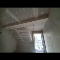 GEFEST IVAN BRAZHNYK - Sufit podwieszany z płyt gipsowo-kartonowych w trakcie montażu, widoczne łączenia i przewody elektryczne, surowe ściany w szarym odcieniu, okno z widokiem na zewnątrz.