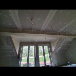 GEFEST IVAN BRAZHNYK - Poddasze w trakcie adaptacji: widoczne płyty gipsowo-kartonowe na skosach ścian i suficie, szpachlowane łączenia, wystające przewody elektryczne, okna dachowe z widokiem na zewnątrz.