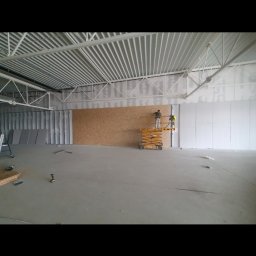 GEFEST IVAN BRAZHNYK - Pracownik na podnośniku montuje płyty w dużym, surowym wnętrzu hali, widoczne profile konstrukcyjne pod sufitem i przygotowane płyty na podłodze.