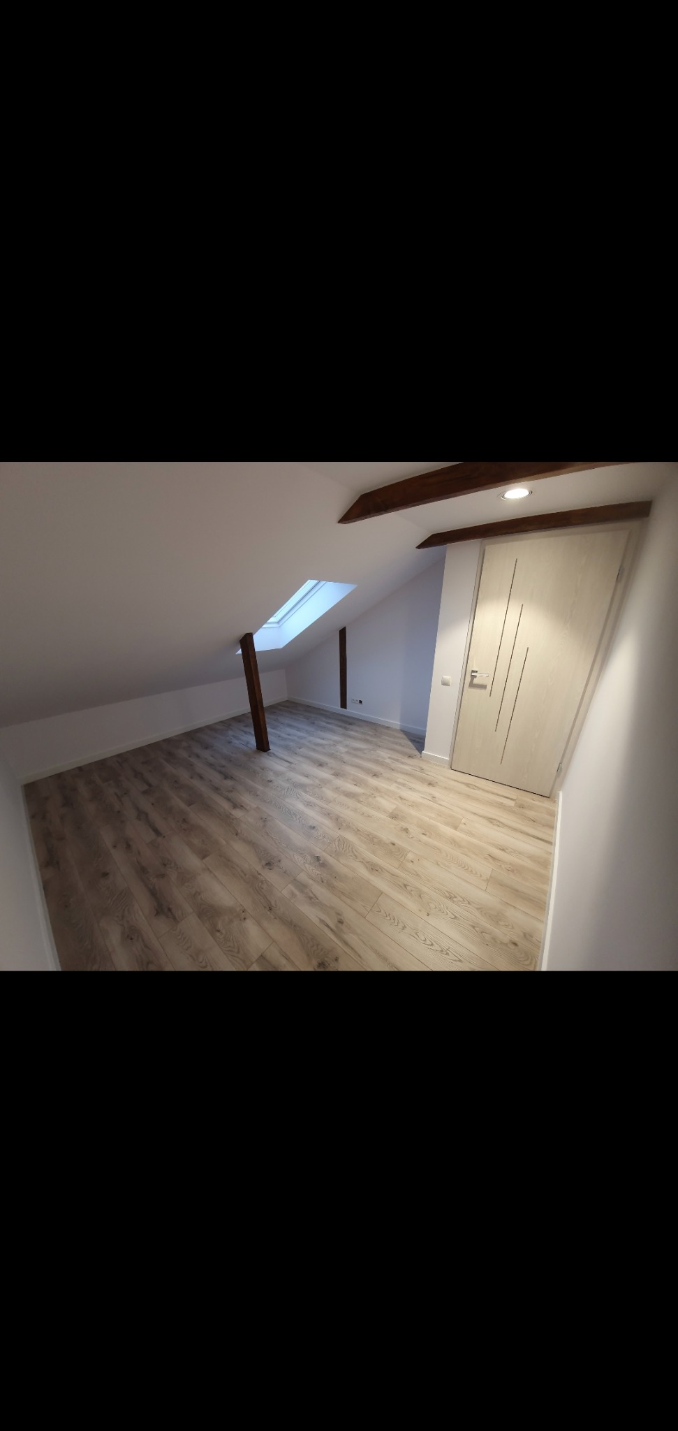 Poddasze po remoncie z drewnianą podłogą, oknem dachowym i drewnianymi belkami stropowymi, białe ściany i drzwi z pionowymi frezowaniami.