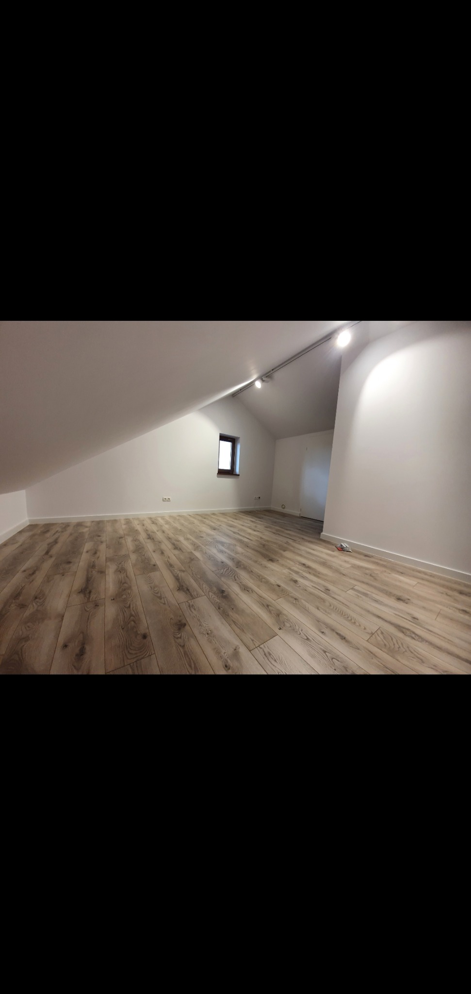 Poddasze po remoncie: jasne panele podłogowe z widoczną strukturą drewna, białe ściany i sufit ze skosem, małe okno w ciemnej ramie, oświetlenie szynowe.