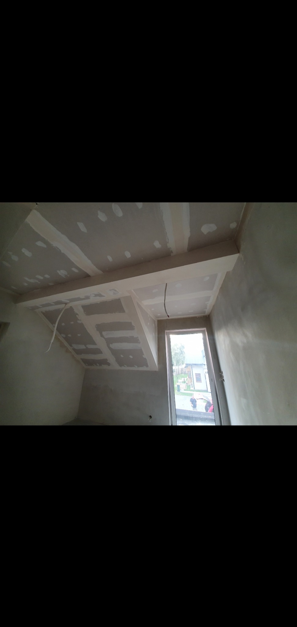 Sufit podwieszany z płyt gipsowo-kartonowych w trakcie montażu, widoczne łączenia i przewody elektryczne, surowe ściany w szarym odcieniu, okno z widokiem na zewnątrz.