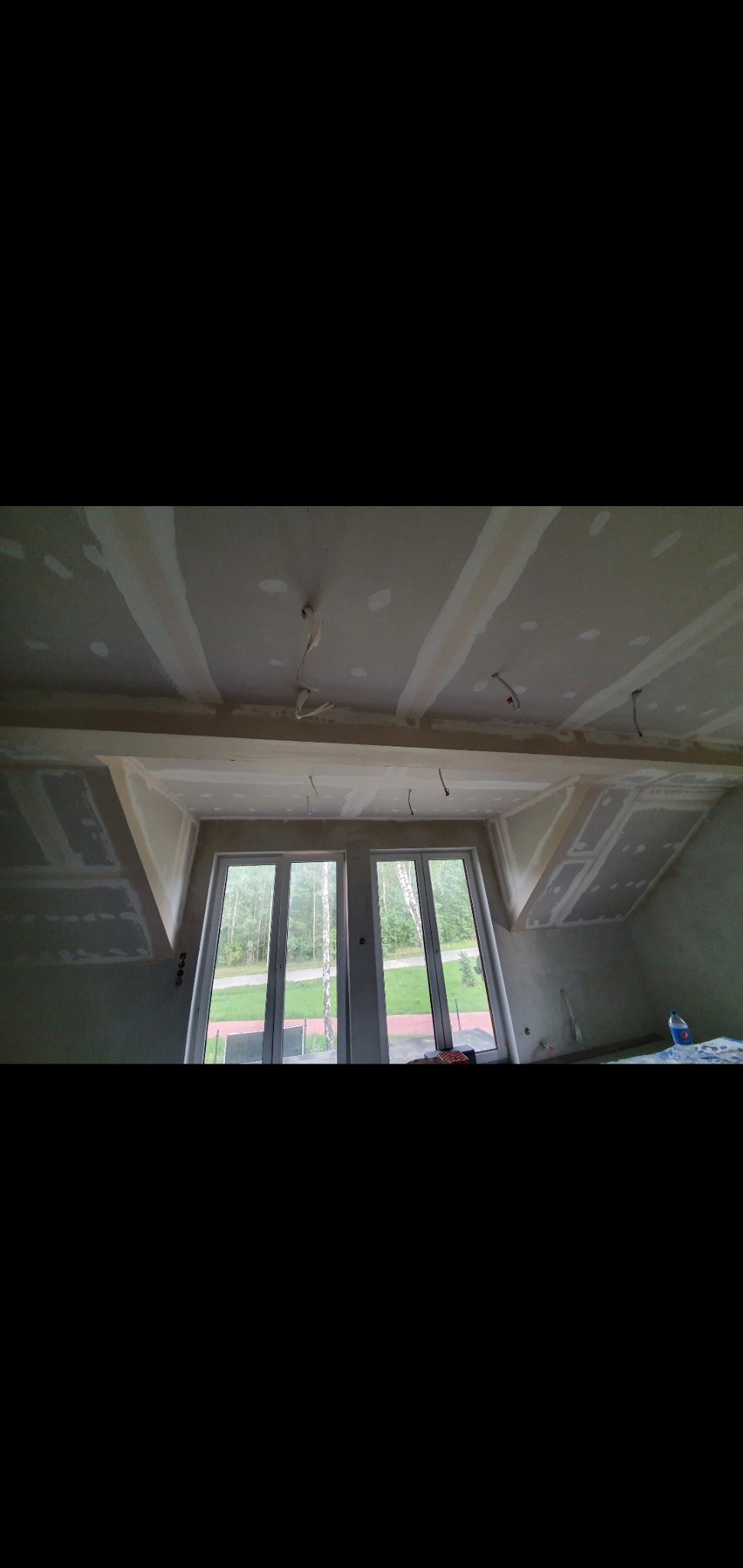 Poddasze w trakcie adaptacji: widoczne płyty gipsowo-kartonowe na skosach ścian i suficie, szpachlowane łączenia, wystające przewody elektryczne, okna dachowe z widokiem na zewnątrz.