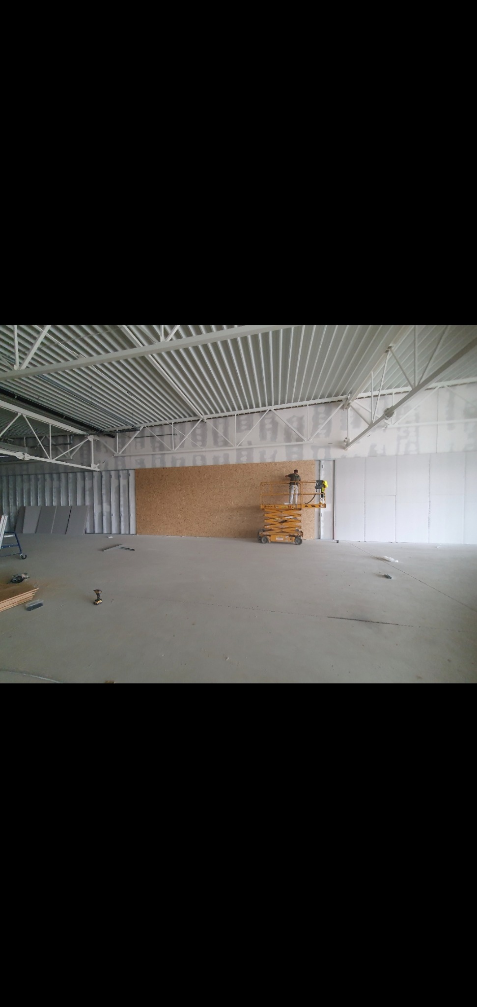 Pracownik na podnośniku montuje płyty w dużym, surowym wnętrzu hali, widoczne profile konstrukcyjne pod sufitem i przygotowane płyty na podłodze.