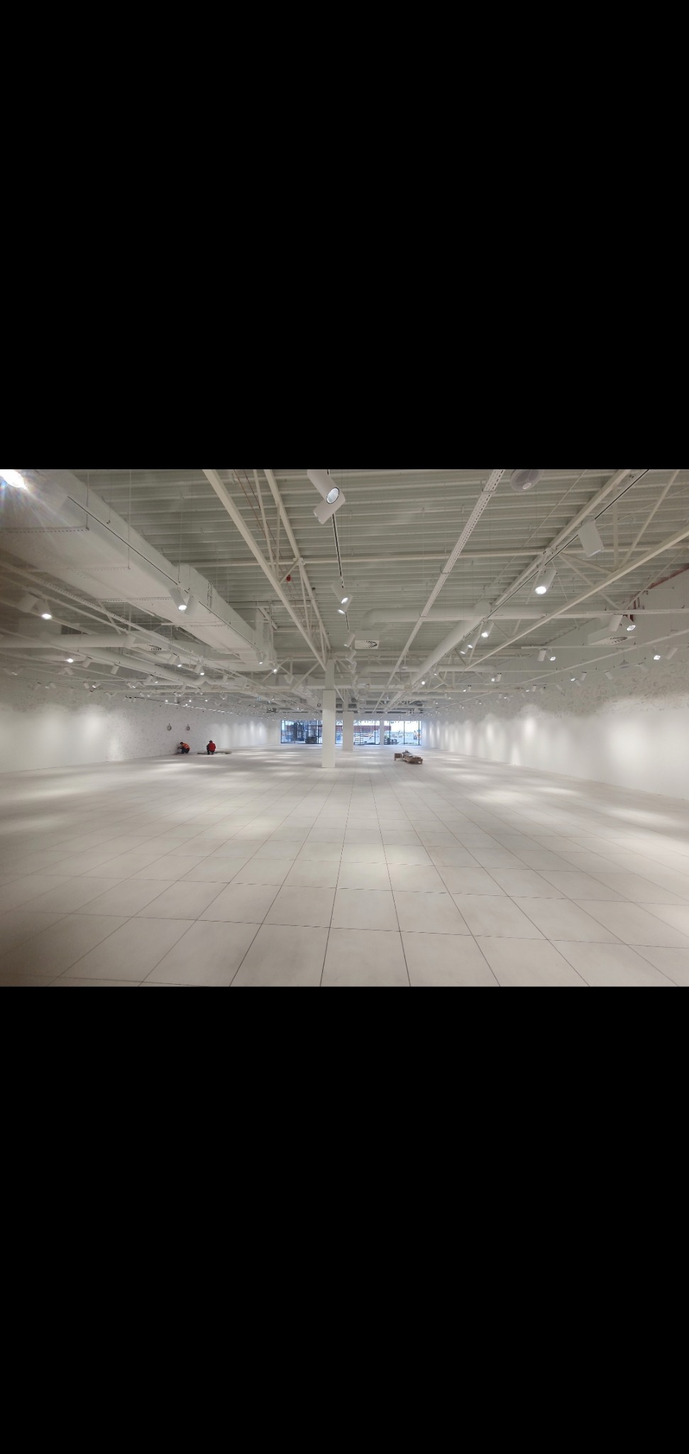 Duża, pusta przestrzeń biurowa z białymi ścianami, podłogą z płytek i metalową konstrukcją sufitową z oświetleniem punktowym. Dwóch pracowników w oddali, przygotowujących podłoże. Świeżo wykończone...