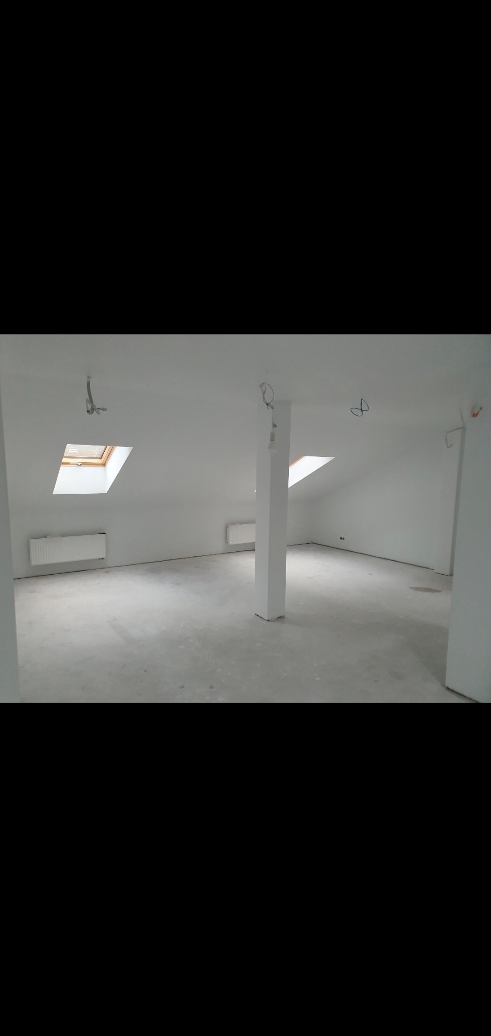 Puste, jasne poddasze po remoncie, z białymi ścianami, kolumną nośną, grzejnikami i oknami dachowymi, przygotowane do dalszych prac wykończeniowych.