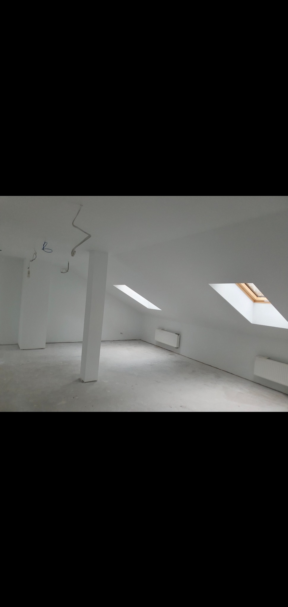 Poddasze po malowaniu na biało, widoczne dwa okna dachowe, grzejniki, słupy konstrukcyjne i wystające kable elektryczne.