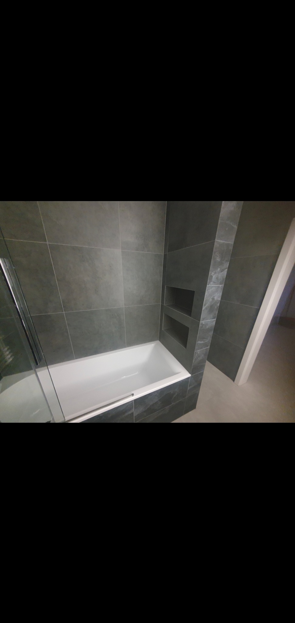 Nowoczesna łazienka z wanną obudowaną szarymi płytkami imitującymi kamień, wbudowane półki w ścianie, kabina prysznicowa z widocznym fragmentem szyby.