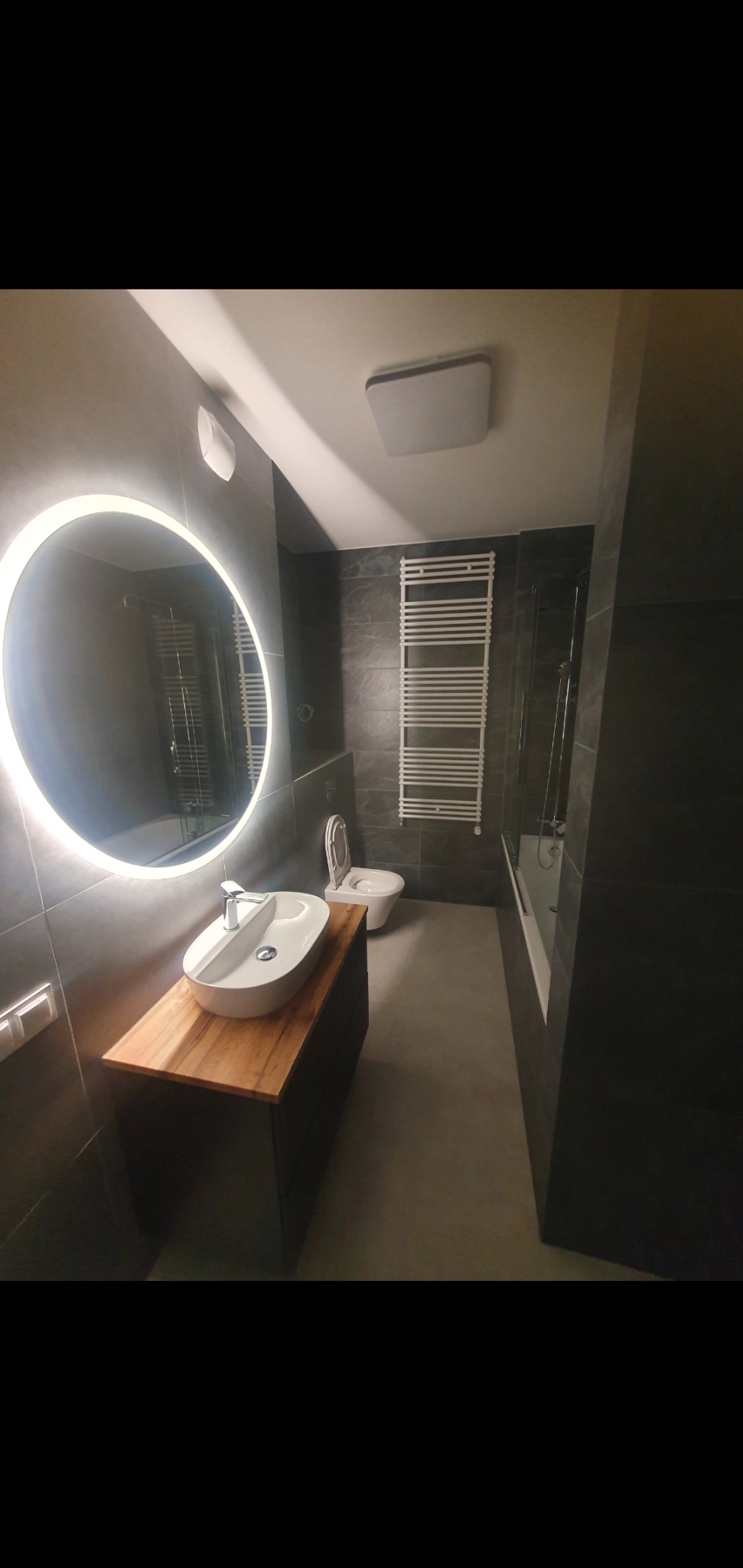 Nowoczesna łazienka z szarymi płytkami, owalną umywalką nablatową na drewnianym blacie, okrągłym lustrem z podświetleniem LED, grzejnikiem drabinkowym i podwieszaną toaletą.