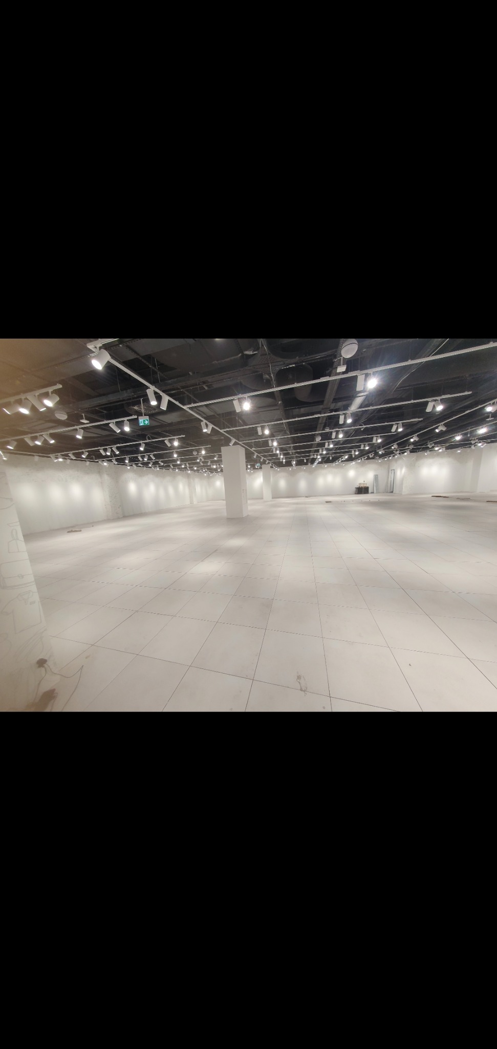 Przestronne, puste wnętrze biurowe po remoncie, z jasnymi ścianami, kafelkową podłogą i nowoczesnym oświetleniem punktowym na suficie. Widoczny filar i ślady po pracach wykończeniowych w rogu.