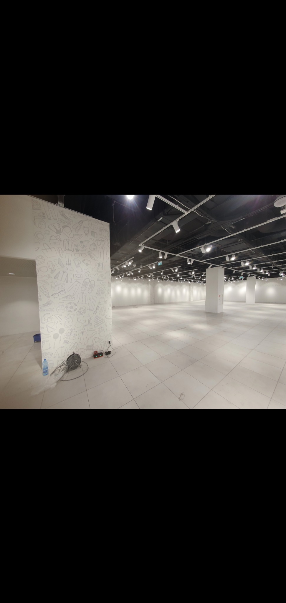 Wnętrze pustego lokalu handlowego z białymi płytkami na podłodze, surowym sufitem z oświetleniem punktowym i ścianą z rysunkami ubrań, butów i akcesoriów, widoczne narzędzia i butelka wody.