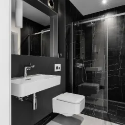 Nowoczesna łazienka z czarnymi marmurowymi ścianami, białą wiszącą umywalką i toaletą, kabiną prysznicową ze szklanymi drzwiami przesuwnymi oraz oświetleniem punktowym w suficie.