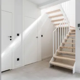 Jasne wnętrze przedpokoju z drewnianymi schodami, białymi ścianami, dwiema parami drzwi z czarnymi klamkami i szarą podłogą z płytek; na ścianie widoczny panel sterujący.