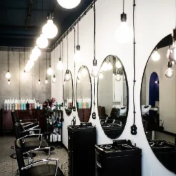 Wnętrze salonu fryzjerskiego z rzędem okrągłych luster, czarnymi fotelami fryzjerskimi, mobilnymi szafkami na kółkach i dekoracyjnymi żarówkami Edison zwisającymi na czarnych kablach.