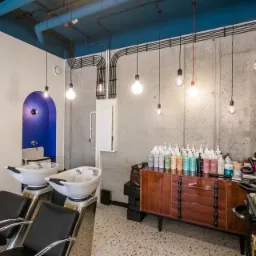 Wnętrze salonu fryzjerskiego z dwoma stanowiskami do mycia włosów, betonową ścianą, wiszącymi żarówkami i szafką z kosmetykami, utrzymane w stylu industrialnym z akcentami kolorystycznymi.